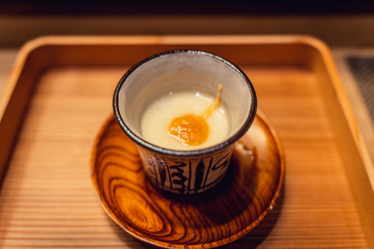 木の盆に置かれた陶製の湯のみに入った薄黄色の出汁の写真